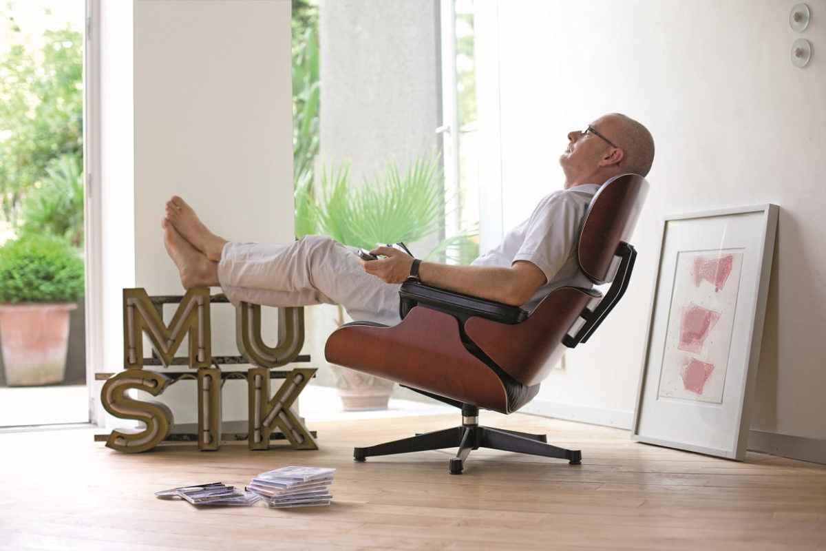 Mann mit Brille sitzt entspannt in einem Sessel und legt seine Füße auf einem Musikschriftzug ab während er Musik hört