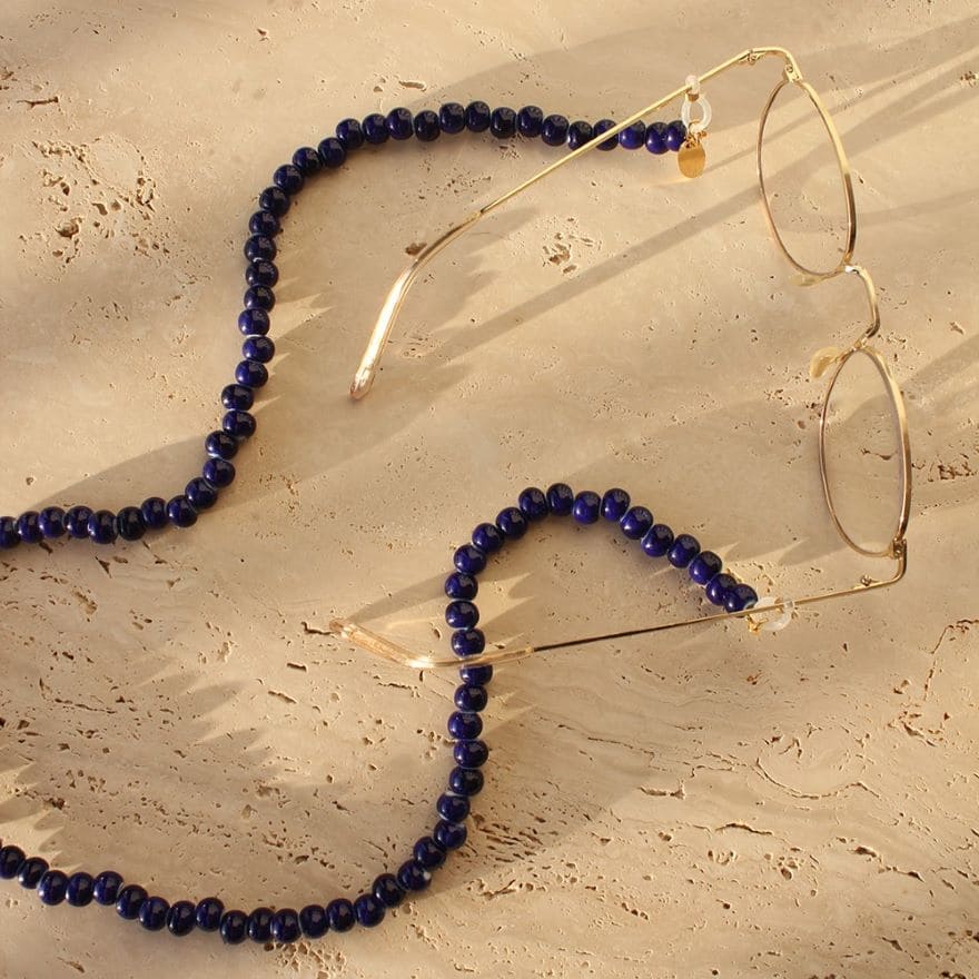 Brillenkette Azul Ceramico von Cocobonito Sunnychords auf einer beigen Steinplatte mit goldfarbener Metallbrille