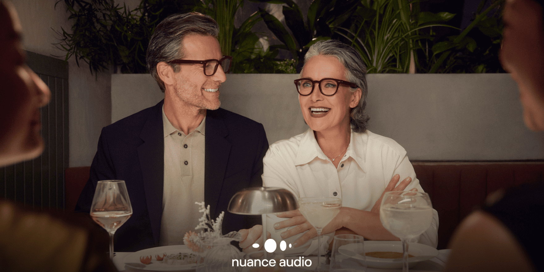 Paar in lustiger Runde im Restaurant. Beide tragen Hörbrille Nuance Audio von GRONDE Sehen & Hören