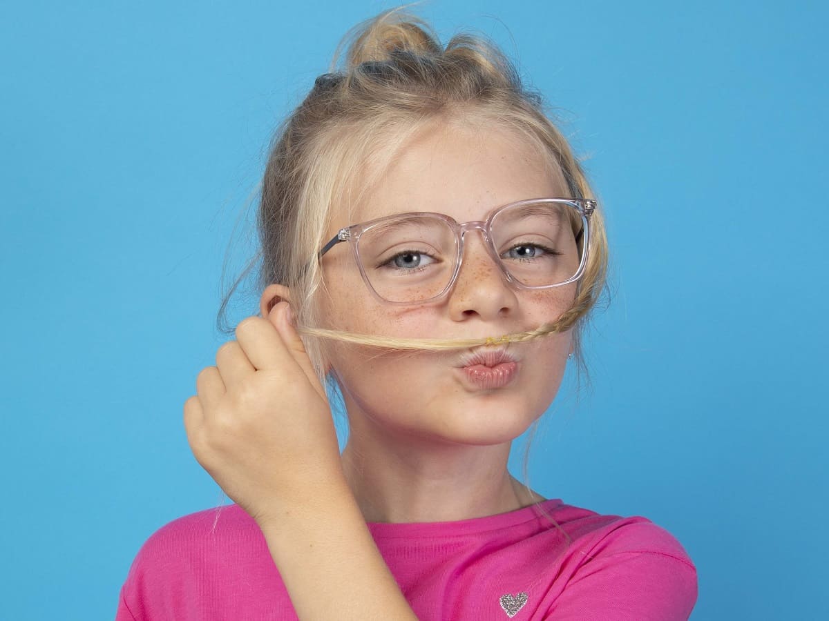 Mädchen in pink trägt Kinderbrille und legt sich eine Haarsträhne als Schnurrbart auf die Lippen