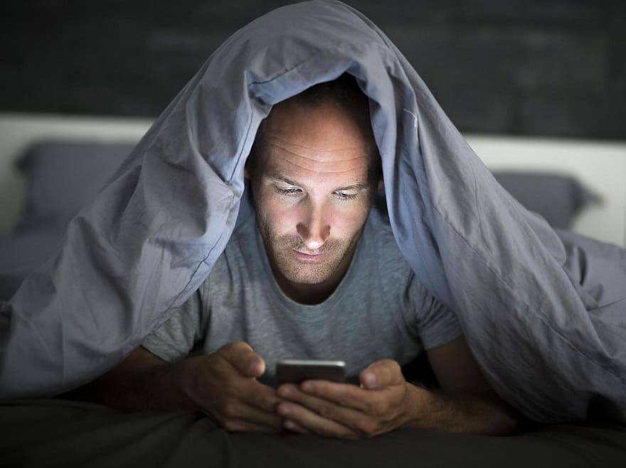 Blaulichtfilter-Brille? Jetzt lesen im Blog von GRONDE Sehen & Hören Mann mit Smartphone unter Bettdecke. Leider ohne Brille mit Blaulichtfilter von Optiker Gronde